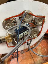 Close-up of a guitar's tremolo system mounted on a white headstock, featuring silver metal potentiometers, a blue coil, orange wire, and gray cables on a workshop surface.