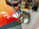 A close-up shot of a vintage silver metal electrolytic capacitor with orange and blue wire leads, mounted on a white circuit board next to gold-colored solder pads and other electronic components.