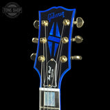 Close-up of a Gibson electric guitar headstock featuring a vibrant blue and black design with gold tuning pegs and strings, set against a solid black background.