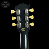 Close-up of the headstock of a Gibson electric guitar against a black background, showing six gold tuning pegs, metal tuning machines, and the Gibson logo in gold on the black backplate.