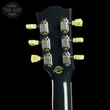Close-up of the headstock of a Gibson electric guitar against a black background, showing six light yellow tuning pegs, silver tuning machines, and the Gibson logo in gold on the black neck.