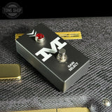 A black metal guitar effect pedal labeled 'M' with a red jewel switch and silver foot switch, resting on a textured gray surface with gold-toned accessories nearby.