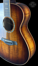 Close-up of an acoustic guitar body showing a rich sunburst finish, natural wood grain, and a black bridge against a black background, with a 'TONG SHOP' watermark in the top right.