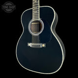 A black acoustic guitar with a glossy finish, white decorative binding, and gold-toned hardware, shown against a solid black background with a Tone Shop watermark in the top-left corner.