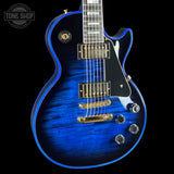 A close-up of a blue electric guitar body with a natural wood grain finish, gold hardware, and a black pickguard, shown against a black background with a Tone Shop watermark in the top left.