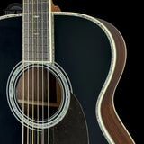 Close-up of an acoustic guitar body showing a glossy black finish, natural wood soundboard edge, decorative rosette, and part of the fretboard against a black background.