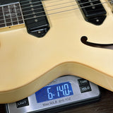 Close-up of a cream-colored electric guitar body resting on a digital scale displaying 6.140 oz, with black pickups and part of the fretboard visible.