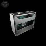 A black textured guitar amplifier cabinet with a clear front window showing a green speaker cone inside, photographed on a solid black background. A Tone Shop watermark is visible in the top-left corner.