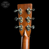 Close-up of the headstock of an acoustic guitar, showing six tuning pegs and a warm brown wood finish against a solid black background. A Tone Shop watermark is visible in the top-left corner.