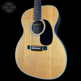 An acoustic guitar with a natural wood finish and black pickguard, shown against a solid black background with a Tone Shop watermark in the top-left corner.