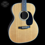 A close-up studio shot of an acoustic guitar body against a solid black background, featuring a natural wood grain finish, a black pickguard, and a white binding. A 'TONE SHOP' watermark is visible in the top-left corner.