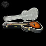 An acoustic guitar with a sunburst finish resting inside an open black hard case with gray padding, shown against a solid black background. A Tone Shop logo watermark is visible in the top-left corner.