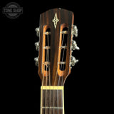 Close-up of the headstock of an acoustic guitar against a black background, showing six silver tuning pegs, two sound holes, and a gold-toned logo inlay. A Tone Shop watermark is visible in the top-left corner.