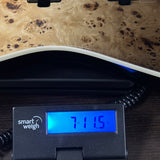 A close-up of a digital smart weigh scale displaying 71.15 kg, positioned underneath a piece of wood with a prominent wavy white edge and a natural burl wood grain top.