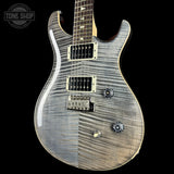 A close-up of an electric guitar body with a silver-gray flamed maple finish, featuring dual humbucker pickups, a bridge, and control knobs, set against a solid black background.