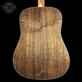 Close-up of the back of an acoustic guitar showing a rich walnut wood grain finish, set against a solid black background with a Tone Shop logo in the top left corner.