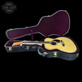 An acoustic guitar with a natural wood top and dark neck, displayed inside an open black hard case with a burgundy interior, set against a solid black background. A Tone Shop logo is visible in the top-left corner.