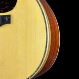 Close-up detail of an acoustic guitar body showing the natural maple top, a dark brown wood side edge, a black pickguard, and a decorative inlay against a black background.