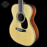 An acoustic guitar with a natural light-colored spruce top and a reddish-brown wooden side, shown against a solid black background. A Tone Shop logo is visible in the top-left corner.