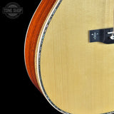 Close-up of an acoustic guitar body showing the light natural wood top, the reddish-brown wood edge, and part of the black bridge against a black background.