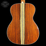 Close-up of the back of an acoustic guitar showing two rich wood grain panels separated by a light natural inlay, set against a solid black background with a Tone Shop logo in the top-left corner.