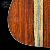 Close-up detail of an acoustic guitar body showing rich reddish-brown wood grain, a light natural wood inlay, and a white edge, with a Tone Shop watermark in the top-left corner.