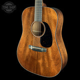A close-up studio shot of an acoustic guitar body against a black background, showing a warm medium-brown wood grain finish, a black pickguard, and a silver bridge. A Tone Shop watermark is visible in the top-left corner.