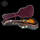 An acoustic guitar with a sunburst finish displayed inside an open black hard case with a burgundy velvet interior, set against a solid black background.