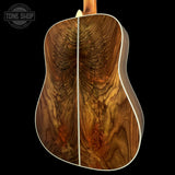 The back of an acoustic guitar featuring a highly figured wood top with dramatic wavy grain patterns in rich brown and amber tones, finished with a white binding edge, shown against a solid black background.