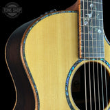 Close-up of an acoustic guitar body showing the natural wood grain top, sound hole rosette with blue inlay, dark sides, and part of the fretboard with floral inlays. A Tone Shop watermark is visible in the top left.