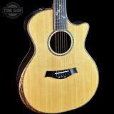 A close-up of a Taylor acoustic-electric guitar body with a natural wood finish, featuring a cutaway top, soundhole, and black bridge against a black background.