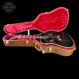 An acoustic guitar with a glossy black body and floral inlay, displayed inside an open brown hard case with bright pink velvet lining, set against a solid black background.