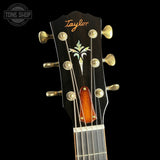 Close-up of a Taylor acoustic guitar headstock against a black background, showing six gold tuning pegs, a palm tree inlay, and an orange wood bridge.