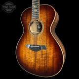 A close-up studio shot of an acoustic guitar body against a black background, showing a warm brown sunburst wood finish with visible grain. A circular Tone Shop watermark is visible in the upper left corner.