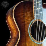 Close-up of an acoustic guitar body showing a rich brown wood grain finish, the sound hole with a blue inlay, and part of the dark fretboard against a black background. A Tone Shop watermark is visible in the top left corner.