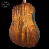 Close-up of the back of an acoustic guitar showing a rich, warm brown wood grain finish, set against a solid black background with a Tone Shop watermark in the top-left corner.