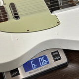 A close-up of a white electric guitar body resting on a digital scale displaying 6.06 lbs, with the guitar's cream pickguard and pickups visible.