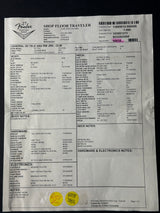 A white paper shop floor traveler form with printed text, barcodes, and handwritten yellow stickers, photographed on a dark surface.