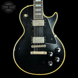 A black electric guitar with a gold outline, featuring a black pickguard, dual humbucker pickups, and four black control knobs, shown against a black background with a Tone Shop logo in the top left corner.