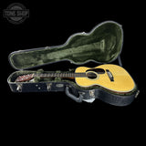 An acoustic guitar with a natural wood body and dark headstock, displayed inside an open black hard case with green velvet lining against a black background. A Tone Shop watermark is visible in the top-left corner.