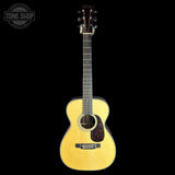 A natural wood acoustic guitar with a cutaway body, shown vertically against a solid black background. A circular 'TONE SHOP' watermark is visible in the top-left corner.
