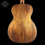 Close-up of the back of an acoustic guitar showing a rich, warm brown wood grain finish against a solid black background, with a Tone Shop watermark in the upper left corner.