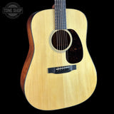 A close-up of a natural wood acoustic guitar body with a dark brown pickguard and black bridge, shown against a solid black background with a Tone Shop watermark in the top-left corner.