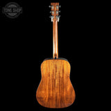 The back of a wooden acoustic guitar, showing the rich grain of the mahogany-style body and neck, set against a solid black background with a Tone Shop logo in the top-left corner.