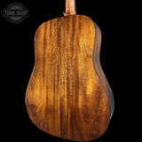 Close-up of the back of an acoustic guitar showing a rich, warm brown wood grain finish, set against a solid black background with a Tone Shop watermark in the top left corner.