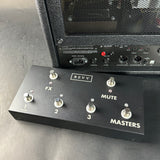 A black REVV guitar effects pedal with five silver footswitches and white labels, positioned in front of a black amplifier on a dark gray surface.