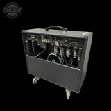 A dark gray guitar amplifier with its front panel removed to show the internal speaker and wiring, featuring a top handle and caster wheels, photographed against a solid black background.