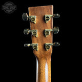 Close-up of the headstock of an acoustic guitar, showing six tuning pegs and a warm brown wood finish against a black background. A Tone Shop logo is visible in the top-left corner.