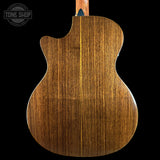 Close-up of the back of an acoustic guitar showing a rich, warm brown wood grain finish, set against a solid black background with a Tone Shop watermark in the top left corner.