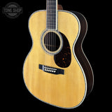 An acoustic guitar with a natural wood finish and dark pickguard, shown against a solid black background with a Tone Shop watermark in the top-left corner.